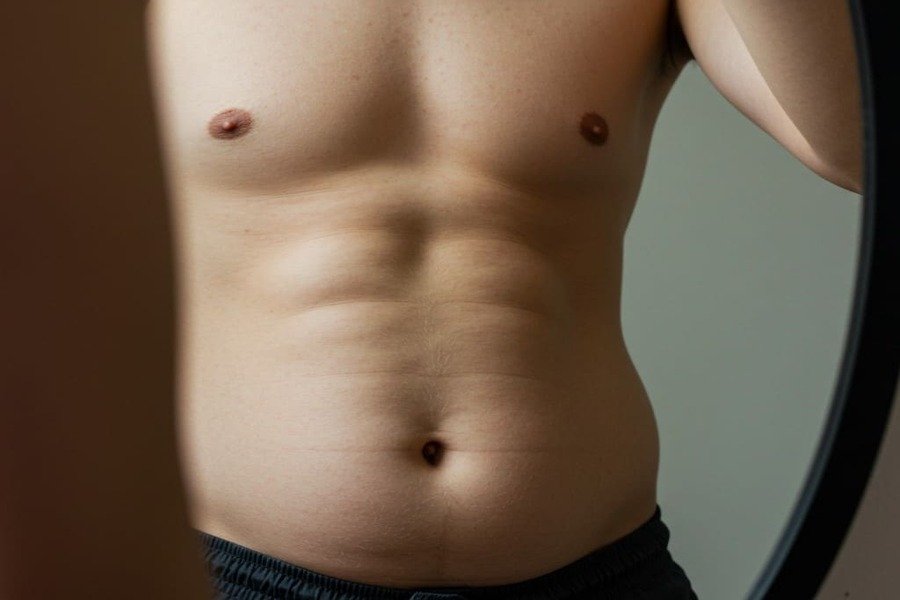 Pessoa olhando no espelho com expressão pensativa, focando na barriga com gordura abdominal visível.