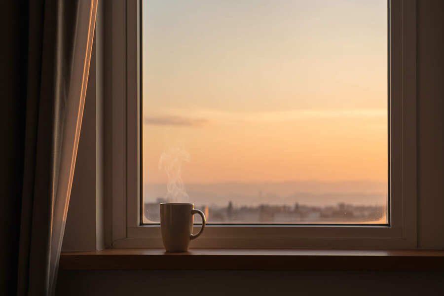 benefícios de acordar cedo. Vista de um amanhecer suave pela janela, com luz natural entrando e uma xícara de café fumegante, simbolizando tranquilidade e um início de dia mais leve.