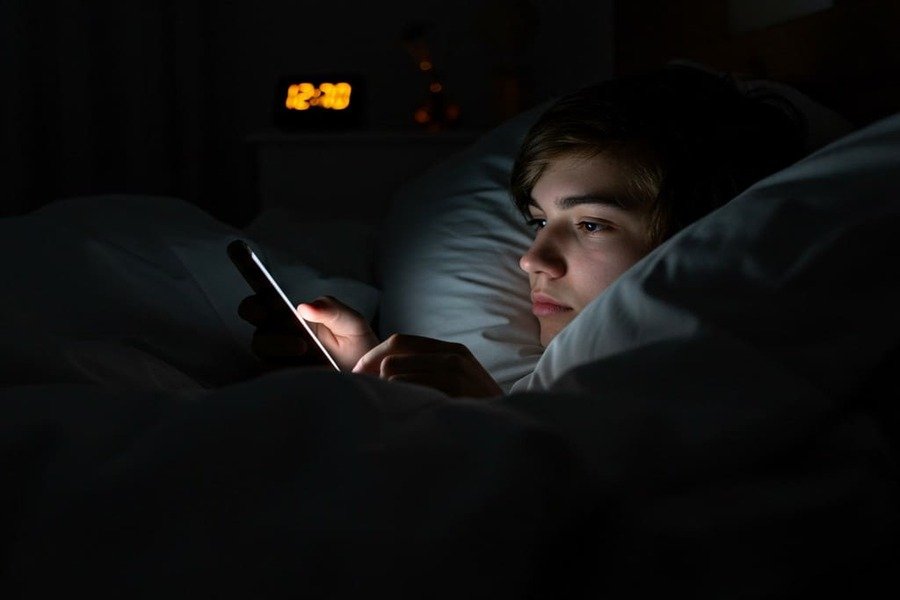 Pessoa deitada na cama à noite olhando para o celular com a luz da tela iluminando o rosto.