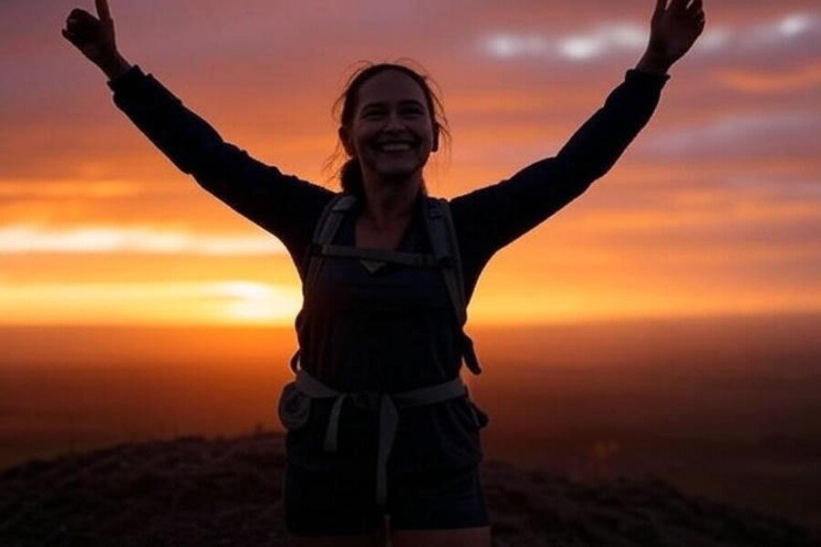 • Mulher com os braços erguidos em comemoração no topo de uma colina ao pôr do sol, simbolizando conquista pessoal após emagrecer.