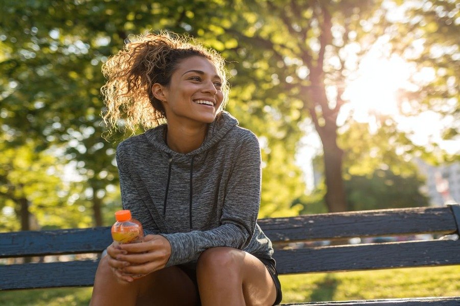 Pessoa sentada em banco de parque após corrida matinal, sorrindo, segurando água e um lanche leve.