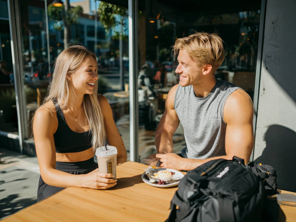 Duas pessoas conversando em uma cafeteria após o treino, uma com shake de proteína e outra com café da manhã leve, representando diferentes abordagens de alimentação.