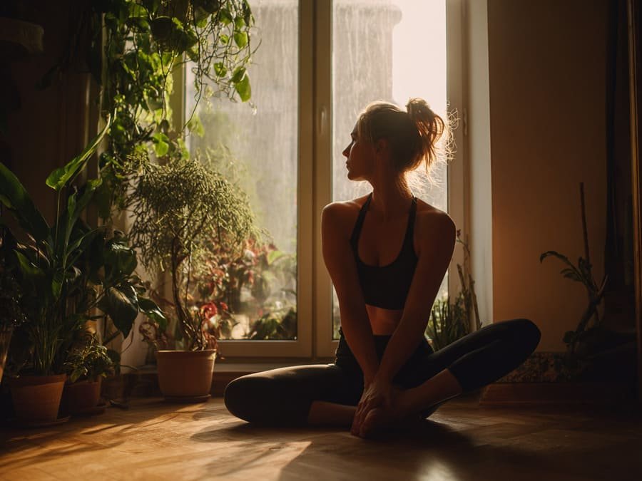 Autocontrole e exercício. Pessoa fazendo alongamento em casa ao lado de plantas, em ambiente calmo e iluminado naturalmente.
