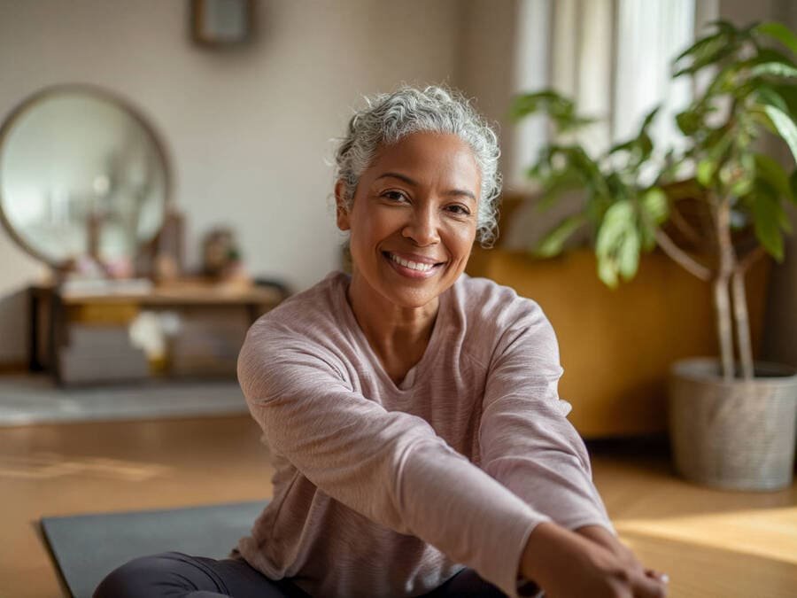 Pessoa sorrindo enquanto se alonga em casa, com tapete de yoga no chão e ambiente acolhedor.