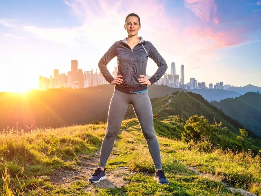 Fotografia de uma pessoa confiante em pé em uma trilha ao entardecer, com braços abertos, céu laranja e rosa ao fundo, simbolizando conquista e transformação.