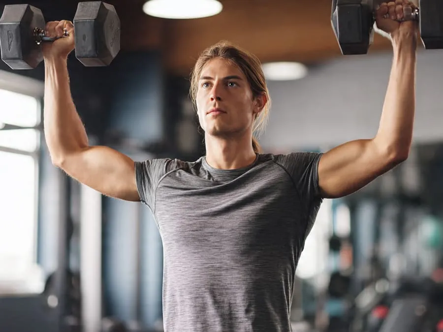 Imagem de um atleta realizando elevação lateral com halteres, destacando os músculos do ombro em uma academia com fundo de pesos e espelhos.