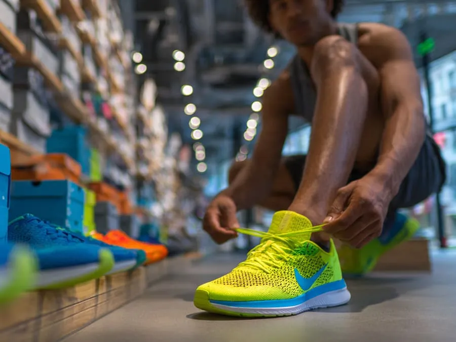 Fotografia de um corredor experimentando tênis de corrida em uma loja esportiva, com caixas de tênis coloridos ao fundo, em um ambiente bem iluminado.