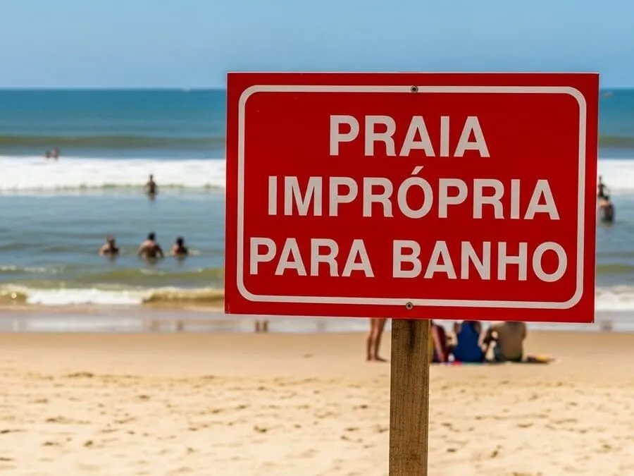 praias impróprias - Fotografia realista de uma placa vermelha de "Praia Imprópria para Banho" fincada na areia de uma praia brasileira, com mar ao fundo.