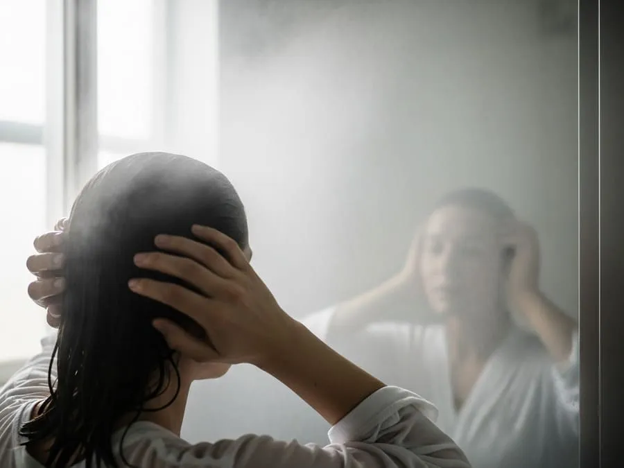 Pessoa de costas em frente a espelho embaçado de banheiro pós-banho, com luz natural suave entrando pela janela.