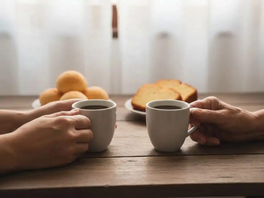 Mãos jovem e idosa segurando xícaras de café sobre mesa brasileira, simbolizando amizade duradoura.