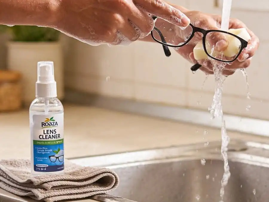 Mãos limpando óculos com água morna e sabão neutro, pano de microfibra ao lado, luz natural, ilustrando método seguro de limpeza.