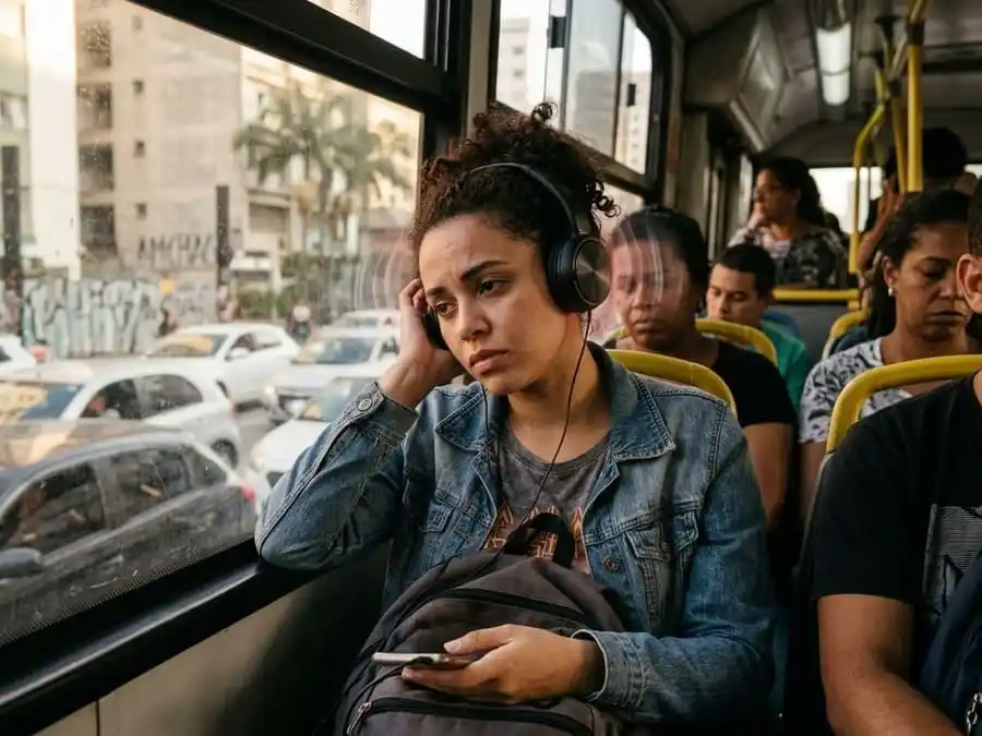 Pessoa usando fone de ouvido com expressão de desconforto, mão no ouvido, ambiente brasileiro cotidiano, luz natural ou de tela, simbolizando o risco cumulativo de uso prolongado e o sinal de alerta do tinnitus.