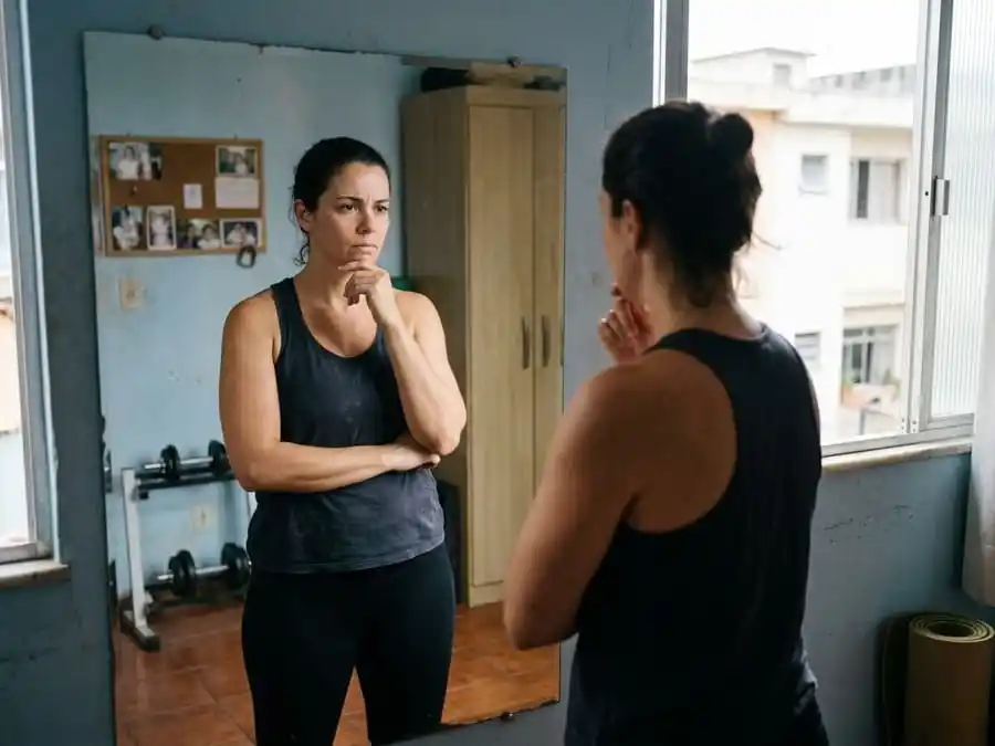 Pessoa olhando para o espelho com expressão pensativa, corpo em transição, quarto ou academia brasileira com luz natural, simbolizando a dúvida entre perda de gordura e ganho de massa muscular.