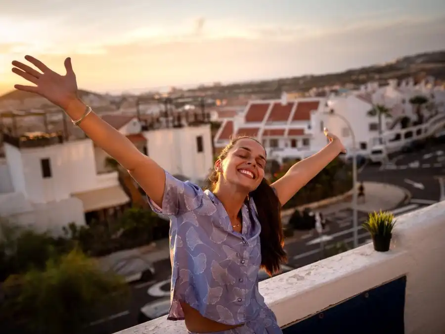 Pessoa sorrindo e dançando com os braços abertos em uma varanda iluminada pela luz dourada do fim da tarde.