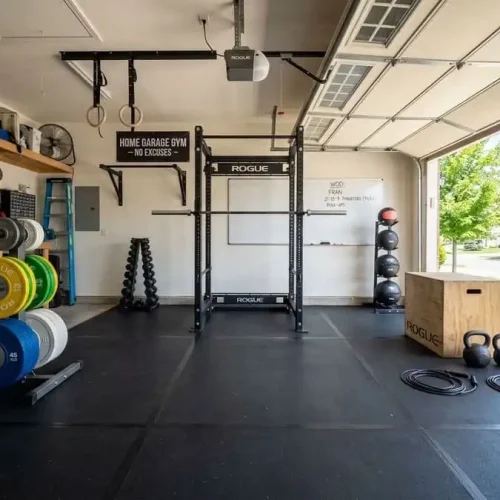 Espaço de treino funcional em uma garagem, com piso de borracha preta, rack de agachamento, barras, anilhas coloridas e acessórios de CrossFit organizados.