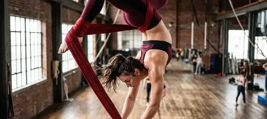 Uma mulher de cabeça para baixo, suspensa por um tecido acrobático vermelho em um estúdio de circo ensolarado, executando um movimento difícil com expressão concentrada.