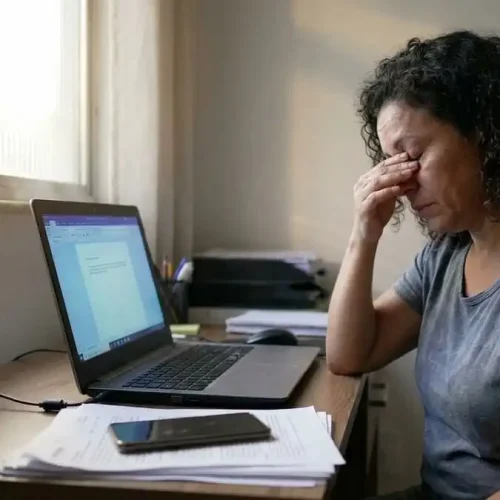 Pessoa com fadiga ocular esfregando os olhos após uso prolongado de tela, luz natural ao fundo, representando o problema comum das telas.