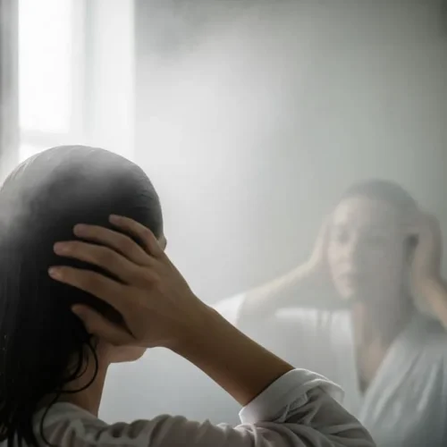 Pessoa de costas em frente a espelho embaçado de banheiro pós-banho, com luz natural suave entrando pela janela.