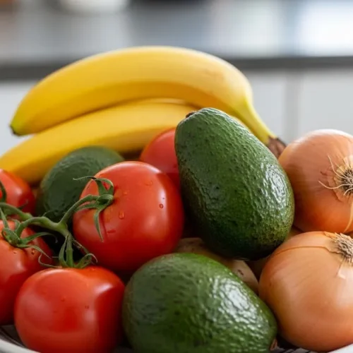 Fotografia de uma fruteira com tomates, bananas, abacates, cebolas e batatas em uma bancada de cozinha, sob luz natural suave.