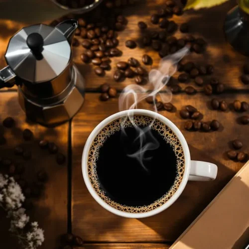 Xícara de café fumegante sobre mesa rústica com grãos de café, cafeteira italiana e caderno ao lado.